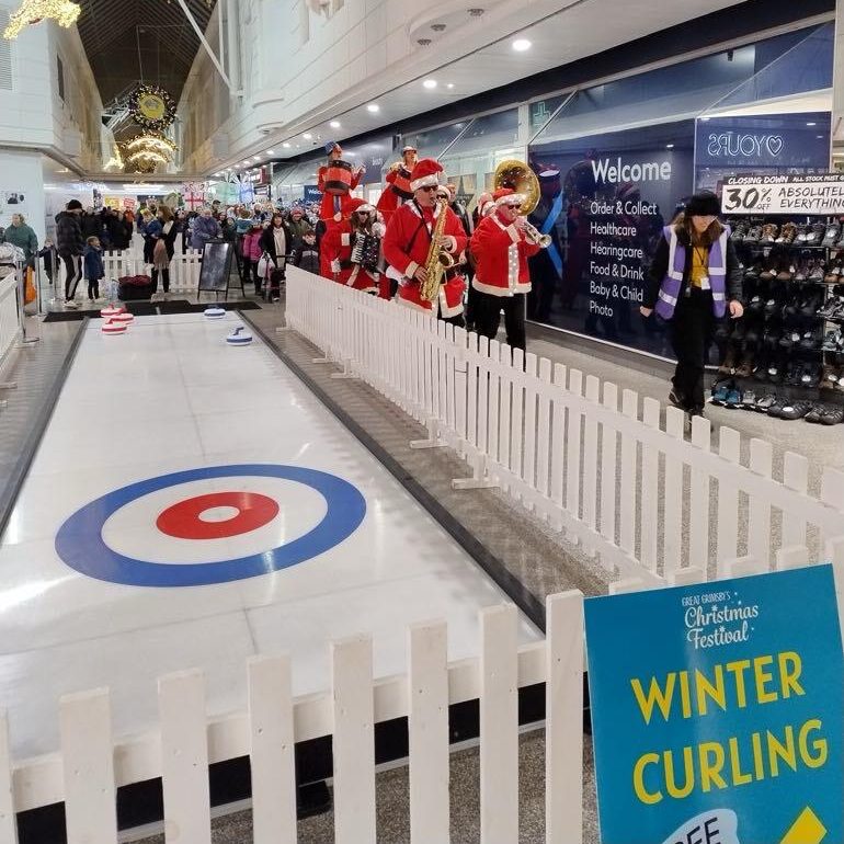 A group of adults dressed up in vibrant Santa suits are playing their band instruments as they walk past a Curling Lane surrounded by a white picket fence, in a shopping centre.