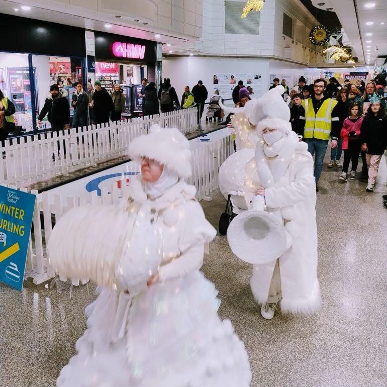 A group of adults dressed up in white Angel outfits are playing their band instruments as they walk past a Curling Lane surrounded by a white picket fence, in a shopping centre.