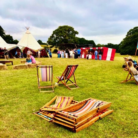 Overview of a Summer event with part of the marquee on the left, our red and white striped Fete Game Side Stalls in a row on the right with deckchairs at the front.