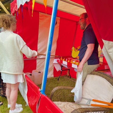 A woman holding one of our Hook a Duck poles, trying to hook a duck, with a member of our staff chatting to her, in our red and white Fete Game Side Stalls.