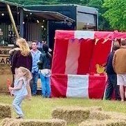Summer event with our hired in red and white striped Fete Game Side Stalls in a row with people watching and playing and a couple of hay bales at the front..