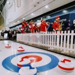A group of adults dressed up in vibrant Santa suits are playing their band instruments as they walk past a Curling Lane surrounded by a white picket fence, in a shopping centre.