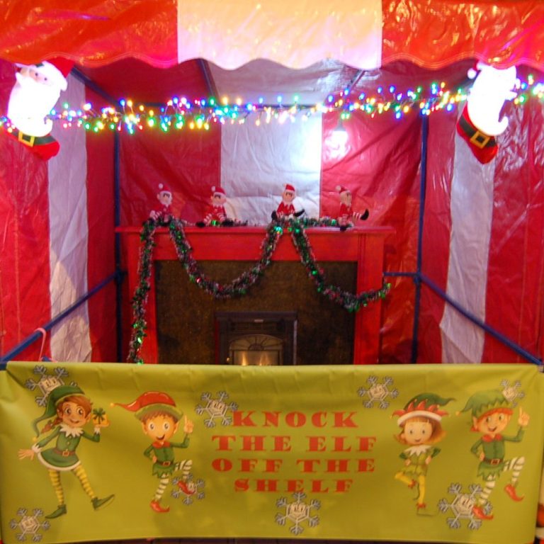 A red and white Carnival Fete Side Stall with 4 soft toy Elves sitting on an actual Fireplace surround ready to be knocked off and a decorated banner at the front of the stall.