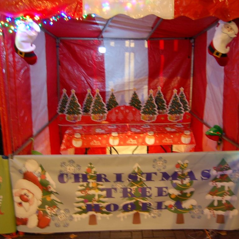 A red and white Carnival Fete Side Stall with Christmas Trees in a group ready for hoops to be thrown over them and a decorated banner at the front of the stall.