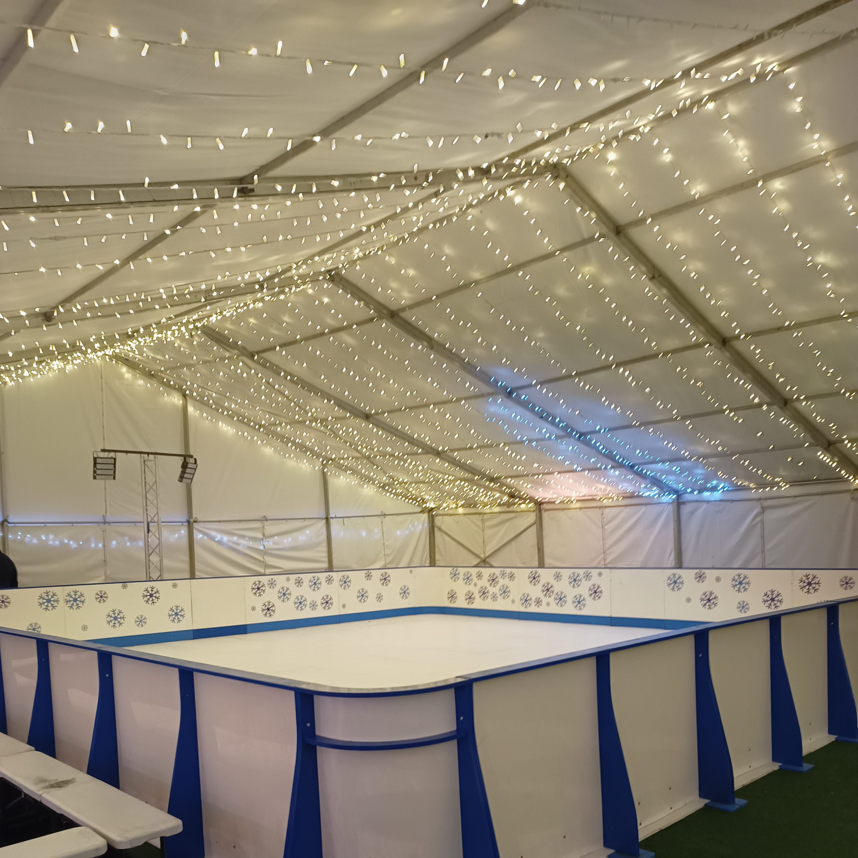 A beautiful festive indoor shot of a hired blue and white Synthetic Ice Rink with bespoke blue snowflake design on the barriers, with hundreds of fairy lights above on the marquee roof.