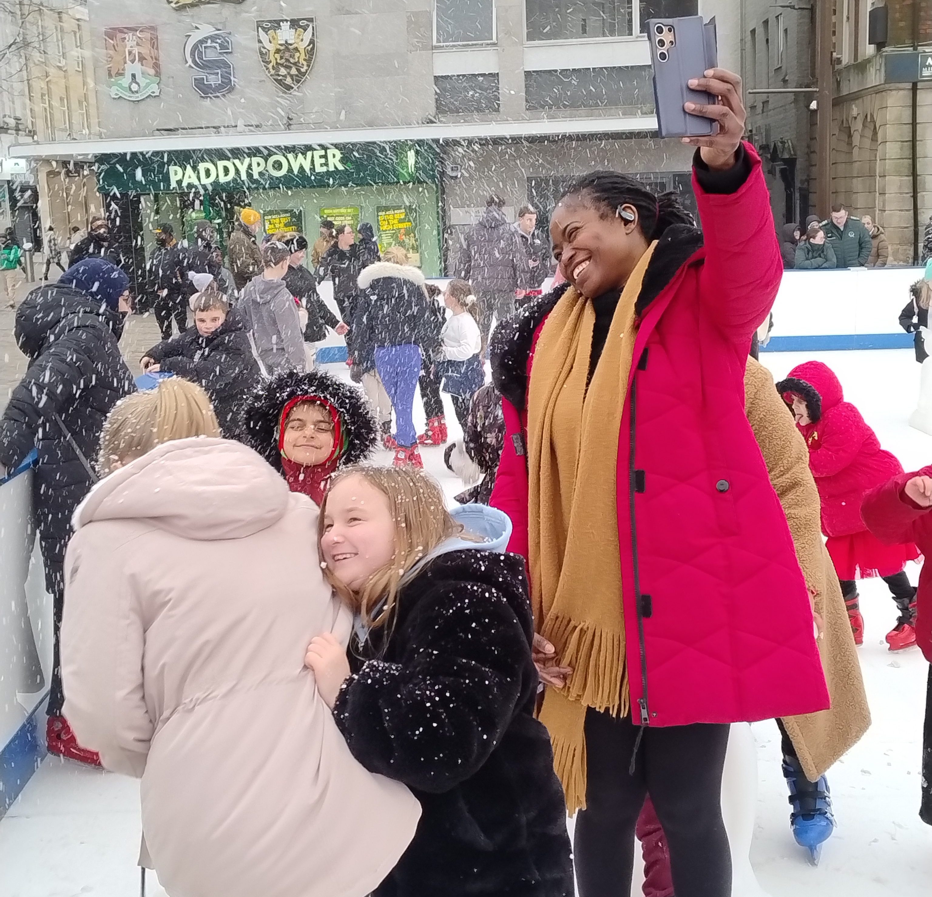A very happy lady in a red coat holding up her phone to capture children on the ice rink being covered by artificial snow.